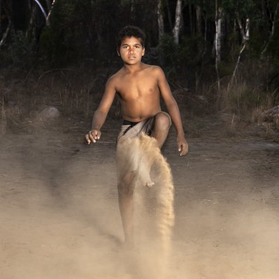 A photograph of boy kicking up sand whilst looking into the lense taken by Mary-Lou Orliyarli Divilli. She is a Nyikina and Ngarinyin woman, living in Kupungarri Community, on the Gibb River Road.